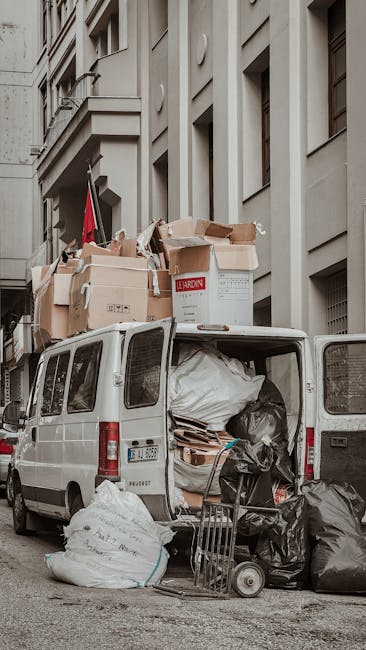 A white cargo van parked on a city street in Mayfair during daylight hours, with its rear doors open revealing the interior filled with various packing materials, including large black and white garbage bags, cardboard boxes, and wooden furniture pieces. The van is loaded with numerous flattened and intact cardboard boxes, some labeled with recycling symbols, along with plastic wrap and packing paper. Several boxes and bags are stacked on the rooftop of the van, secured temporarily for the loading process. An additional trolley and a small hand truck are positioned nearby on the pavement, indicating ongoing packing and loading activities. The van is situated in front of a classic multi-storey building with large windows and decorative architectural features typical of the area. The environment suggests a home relocation operation, minimally involving man and van services, focused on efficiently managing bulky waste and clutter resulting from a house move in accordance with professional removals standards.