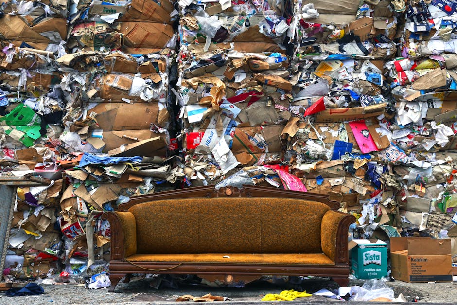A white cargo van parked on a city street in Mayfair during daylight hours, with its rear doors open revealing the interior filled with various packing materials, including large black and white garbage bags, cardboard boxes, and wooden furniture pieces. The van is loaded with numerous flattened and intact cardboard boxes, some labeled with recycling symbols, along with plastic wrap and packing paper. Several boxes and bags are stacked on the rooftop of the van, secured temporarily for the loading process. An additional trolley and a small hand truck are positioned nearby on the pavement, indicating ongoing packing and loading activities. The van is situated in front of a classic multi-storey building with large windows and decorative architectural features typical of the area. The environment suggests a home relocation operation, minimally involving man and van services, focused on efficiently managing bulky waste and clutter resulting from a house move in accordance with professional removals standards.