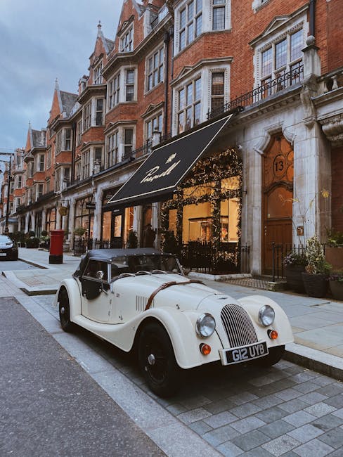 A vintage cream-colored convertible car with a black soft top is parked on a narrow street in Mayfair, London, in front of a row of historic red brick townhouses with detailed stone facades and large sash windows. The car's front grille and round headlights are prominent, and it has a British registration plate reading 'G12 UVB.' Directly behind the car, there is a black awning with the word 'The' partially visible, attached to a building that appears to function as a shop or restaurant. The sidewalk features stone paving, with some potted plants near the entrances of the buildings. The street scene is lit by natural daylight, suggesting a clear or partly cloudy day. In the background, more multi-storey residential buildings with decorative architectural elements are visible. Occasionally, Man and a Van Mayfair's removals services are involved in home relocations and furniture transport along this street, supporting the packing and moving process as part of a professional house removal operation.