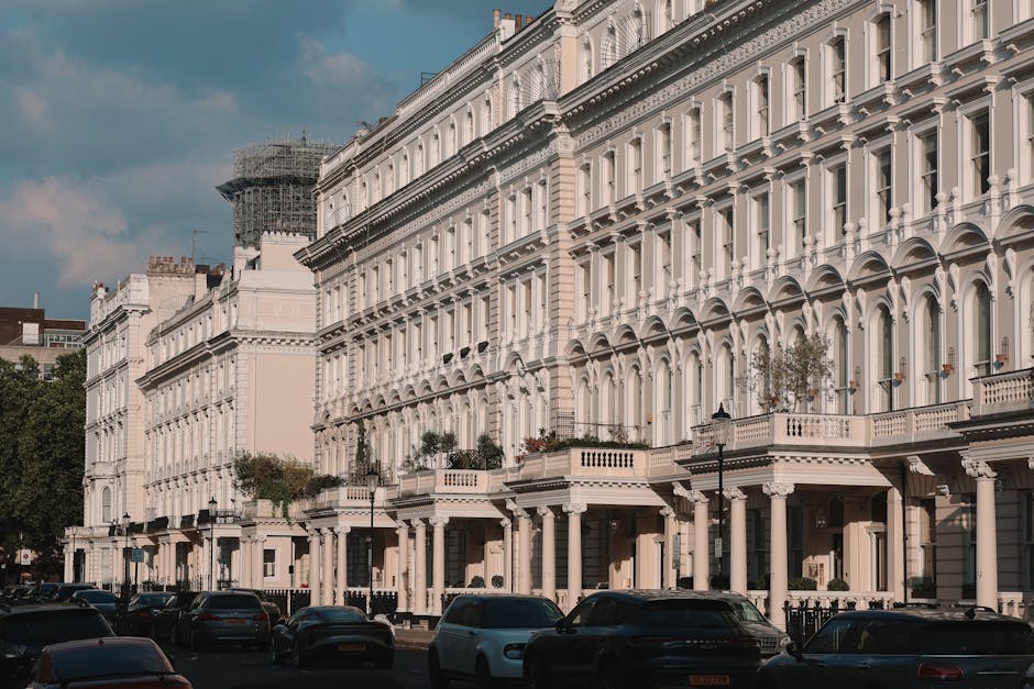 An exterior view of a row of white, multi-story Victorian-style terraced houses with detailed cornices, arched windows, and decorative balconies. The buildings are situated along a street with parked cars lining the pavement. The image is taken during daylight with natural lighting illuminating the facades. Visible on the street level are the entrances to the houses, some featuring small front gardens or potted plants, and a black lamppost is positioned near the sidewalk. In the background, a building under scaffolding suggests ongoing renovation work. This scene depicts an urban residential area, typical of Mayfair properties, and is relevant to house removals, furniture transport, and home relocation services, as provided by [COMPANY_NAME] specializing in removals, with the context of packing and loading processes in a moving operation.