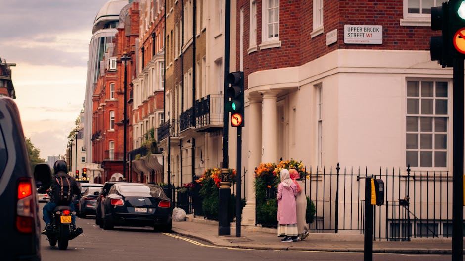 A street scene showing a white moving van parked close to a row of Victorian-style residential buildings with red brick and white facades, large windows, and decorative black balconies, indicative of a typical house removal environment in Mayfair. In the foreground, a person wearing a pink coat and a headscarf stands outside a property, possibly preparing for a home relocation, while a motorcyclist wearing a helmet rides past. The pavement features yellow double lines, black iron railings, and flower arrangements in large planters near the building entrance. Traffic lights are visible, with one showing green, indicating an active loading or moving process. The overcast sky suggests evening or early morning lighting, providing a calm backdrop for furniture transport, packing, and loading activities associated with professional removals by Man and a Van Mayfair.