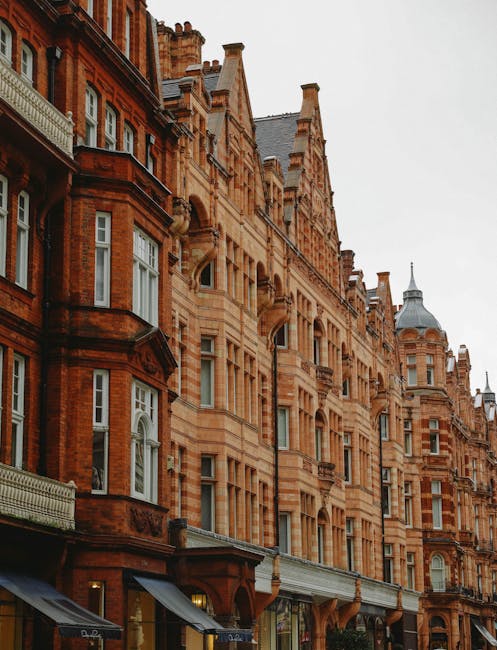 Photograph of a row of historic terraced buildings with ornate red brick facades and decorative architectural details on a street in Mayfair, London. The buildings feature large sash windows, some with white frames, and small balconies with wrought iron railings. The ground floor shops have dark awnings extending over the pavement. In the foreground, a man wearing dark clothing is lifting a cardboard box wrapped in plastic from a pile of similar boxes, which are stacked near the building entrance on the pavement. A transport trolley and moving blankets are partially visible nearby. The scene is set during daylight with overcast weather, indicating an active home relocation or furniture transport process taking place, consistent with professional removals services by Man and a Van Mayfair, capturing a moment of loading or packing effort in preparation for a house move in the Mayfair area.