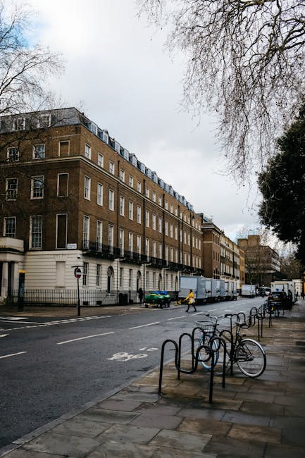 A street scene in an urban area with tall, multi-storey buildings on either side, featuring a small loading zone with two white moving trucks branded with 'uniserve' and 'Becton Transport' parked closely together; a black scooter is parked on the pavement beside the trucks. The pavement is divided by white lines indicating a designated loading area or parking zone. Two pedestrians walk across the street; one is a woman dressed in dark clothing carrying a bag, and the other is an older man with white hair, wearing a dark jacket and trousers. The scene is well-lit with natural daylight, illustrating a typical setting for house relocation or furniture transport, with the duo possibly involved in moving or logistics operations supported by [COMPANY_NAME], as part of a professional removals service near Grosvenor Square, Mayfair.