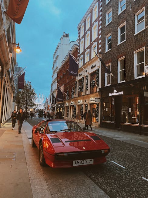 A red classic sports car, parked on a narrow city street with pedestrians walking along the pavement. The street is lined with multi-story buildings featuring various storefronts, some with lit signs, and black flags hanging from the facades. Overhead, decorative street lighting and festive decorations suggest a vibrant urban environment. The scene is captured during twilight, with a blue sky overhead. The surroundings indicate a busy commercial area, typical for high-end districts like New Bond Street and Mayfair W1, where professional removals and relocations by companies like Man and a Van Mayfair are often required for house and office moves involving furniture transport, packing, and loading processes.