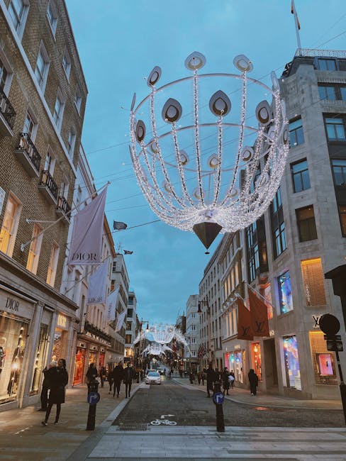 A wide-angle view of a busy shopping street in Mayfair, illuminated by festive decorative chandeliers hanging overhead. The street is lined with luxury retail stores, including Dior and Louis Vuitton, identifiable by their signage and window displays. Pedestrians are walking along the pavement, some in groups and others alone, with a few carrying shopping bags. The scene is set in the late afternoon or early evening, with a clear blue sky and modern buildings on either side. Decorative orb-shaped light fixtures are suspended from the chandeliers beneath the street’s canopy, creating a festive atmosphere. The street is partially closed with bollards and features a designated bicycle lane marked on the pavement. Man and a Van Mayfair’s office relocations and furniture transport services are relevant to this setting, reflecting the logistical coordination involved in moving and packing possessions in an urban shopping environment.