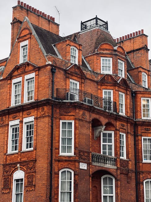 The image shows the exterior of a historic red brick building with multiple floors, featuring large white-framed sash windows and decorative architectural details. On one of the upper balconies, there is a black wrought iron railing, and a small rooftop terrace with a wooden balustrade overlooks the building. The roof has a steep pitch with dormer windows and a chimney visible against a cloudy sky. This type of Victorian-style architecture is typical of Mayfair properties. The building’s exterior appears well-maintained, with clear details of the brickwork and window features. The image captures the grandeur of a classic London townhouse, which may be part of a house removal or relocation project conducted by Man and a Van Mayfair, emphasizing careful handling and preservation of historic features during furniture transport or home relocation activities.