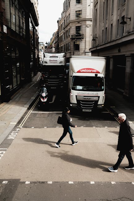 A street scene in Mayfair showing a white delivery van with 'uniserve.co.uk' branding parked partially on the pavement and partially on a road with tram tracks, adjacent to a building with large windows. Two pedestrians, a man and a woman, are crossing the road in front of the van; the woman is dressed in dark clothing and carrying a bag, while the man wears a dark jacket and light trousers. In the background, there is a large white truck with 'Becton Transport' branding and a small motorcycle parked on the pavement nearby. The scene is set in a city environment with multi-storey buildings lining both sides of the street, and the lighting suggests daytime with overcast skies. This image exemplifies urban logistics involving the loading and unloading of furniture or boxes, relevant to house removal and furniture transport services provided by companies like Man and a Van Mayfair.