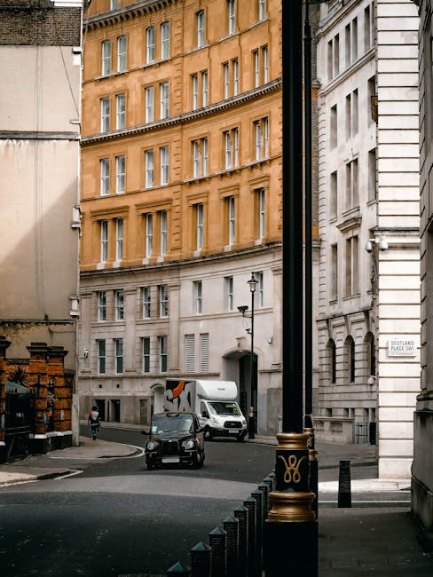 A narrow street in Mayfair with multi-storey buildings featuring orange and white facades, large windows, and architectural details. In the foreground, a black bollard with gold detailing lines the pavement, adjacent to a black pole. Near the corner of the street, a man is walking while wearing casual clothing. A small black car is parked on the road, and a white van is seen further along the street, likely involved in home relocation or furniture transport. Stacked wooden crates and various packing materials are visible on the sidewalk, indicating ongoing packing or loading activities typical of house removals. The scene is lit by natural daylight, with a few street lamps mounted on the buildings. This setting reflects urban logistics connected to rapid moving or clearance services offered by Man and a Van Mayfair, with visible elements of furniture and packing goods prepared for loading into a vehicle.