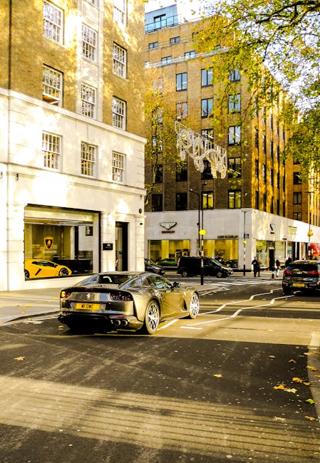 A street scene in Mayfair showing a white delivery van with 'uniserve.co.uk' branding parked partially on the pavement and partially on a road with tram tracks, adjacent to a building with large windows. Two pedestrians, a man and a woman, are crossing the road in front of the van; the woman is dressed in dark clothing and carrying a bag, while the man wears a dark jacket and light trousers. In the background, there is a large white truck with 'Becton Transport' branding and a small motorcycle parked on the pavement nearby. The scene is set in a city environment with multi-storey buildings lining both sides of the street, and the lighting suggests daytime with overcast skies. This image exemplifies urban logistics involving the loading and unloading of furniture or boxes, relevant to house removal and furniture transport services provided by companies like Man and a Van Mayfair.