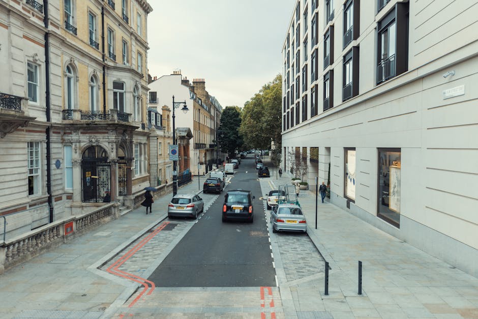 A view of a city street showing a modern white building on the right side with several large windows and a smooth facade, and on the left side, an older ornate building with detailed stonework, balconies, and arched doorways. The street is lined with parked cars, including a grey sedan, a black car, and a white van. In the foreground, a man in casual clothing is walking on the pavement near the older building. The street appears to be part of a home relocation process, with the setting suggesting a professional removals service such as Man and a Van Mayfair. The scene is illuminated by natural daylight with some trees visible in the distance, and the road surface is clean, with designated parking bays and a pavement suitable for loading and unloading large furniture or boxes. The overall environment indicates an urban area prepared for furniture transport or packing and moving activities.