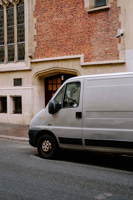 Two movers dressed in white protective suits, face masks, and blue gloves are standing outside of a large white van with its rear doors open, in a parking lot with a clear sky and leafless trees in th