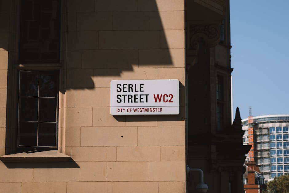 A street sign mounted on the exterior beige stone wall of a building indicating 'Serle Street' in white with black and red lettering, and the code 'WC2' for the postcode district. The sign is positioned beneath a shadow cast by a neighboring structure, with a window visible to the left that has a black metal frame and multiple panes of glass, reflecting the surrounding environment. The scene is lit by natural daylight, with a clear blue sky in the background. In the distance to the right, a modern high-rise building with a curved glass facade is partially visible, contrasting with the historic architecture. This location is relevant to house removals and home relocation services, where accurate street identification is essential for planning moving logistics, including obtaining permits from Westminster City Council, as offered by companies like Man and a Van Mayfair.