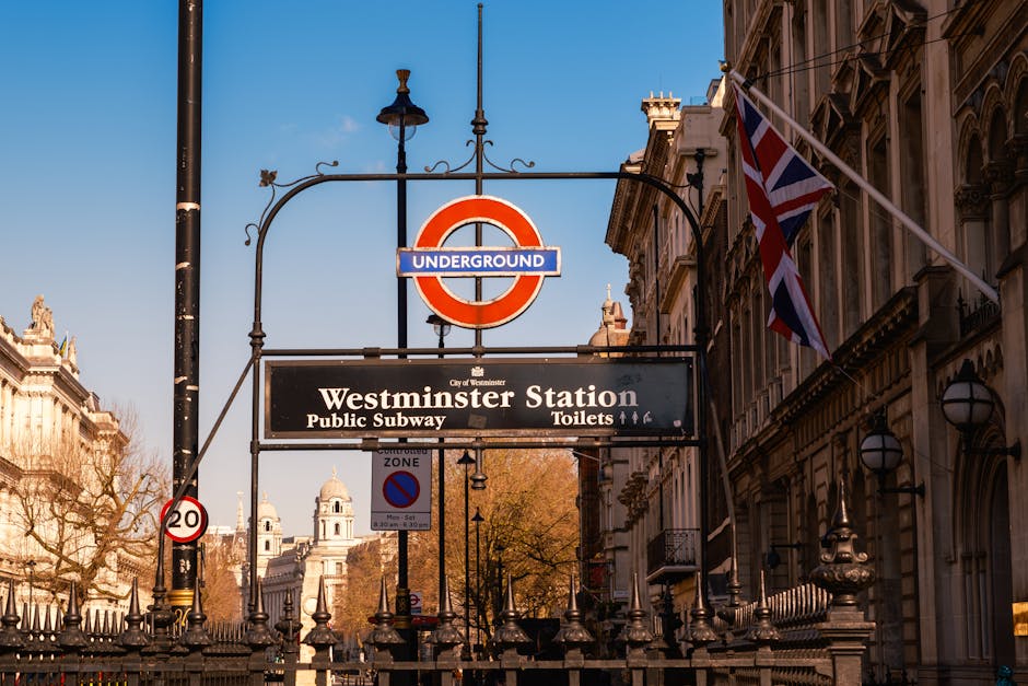 Photograph of Westminster Station in London showing a prominent roundel sign with a blue background and red circle, indicating the underground. Beneath the roundel, there is a black rectangular sign displaying 'Westminster Station' along with information about public subway, toilets, and additional amenities. To the right, a Union Jack flag is attached to a flagpole, fluttering in the wind. The scene is set against historic city buildings with ornate facades illuminated by daylight and a clear blue sky overhead. Street lamps and a lamppost are visible, along with a traffic sign indicating a 20-mph speed limit. The area appears busy with street furniture and elements typical of a central London transport hub, with some trees in the background, suggesting a location suitable for home relocation or furniture transport services, as exemplified by companies like Man and a Van Mayfair.